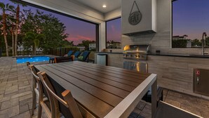 An outdoor kitchen with a grill, a dining table, and a pool in the background.