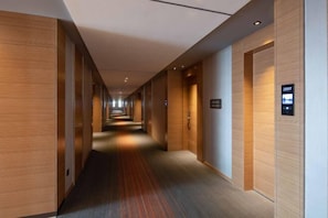 Un pasillo moderno de hotel con paneles de madera, piso alfombrado y un panel digital de ascensor.