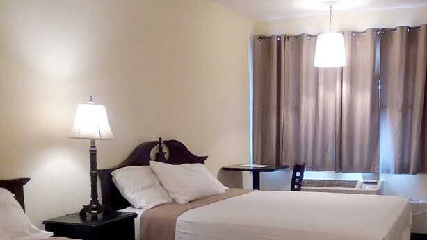 A hotel room with two single beds, a bedside table with a lamp, a window with curtains, and a small table. - Walton Inn (Willard)