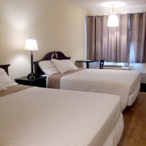 A hotel room with two single beds, a bedside table with a lamp, a window with curtains, and a small table.