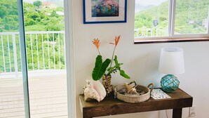 Una habitación con piso de madera, una puerta corrediza de vidrio que da a un balcón, una pequeña mesa con libros, adornos y dos cuadros en la pared.