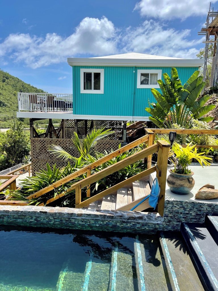 Un edificio turquesa con balcón, piscina con cubierta de madera y plantas tropicales.