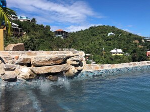 A swimming pool with a rocky edge and a view of a hillside with houses.