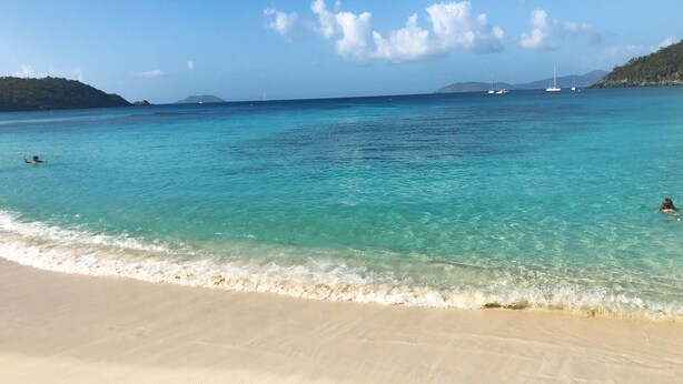 Une plage à l’eau turquoise et limpide, un rivage sablonneux et des bateaux qui se dessinent à l’horizon.