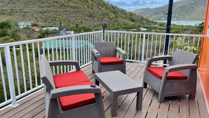Une terrasse dotée de meubles en osier et de coussins rouges, offrant une vue sur un paysage vallonné et une étendue d’eau.