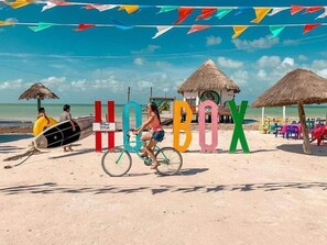 A beach scene with a large 'HOTBOX' sign, a thatched-roof hut, and beach chairs. - Hotel La Casa del Capi   (Isla Holbox)