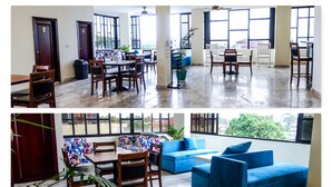 A spacious dining area with wooden tables and chairs, a blue sofa, and large windows offering views of the outdoors. - Sunset Inn Guest House (Monrovia)