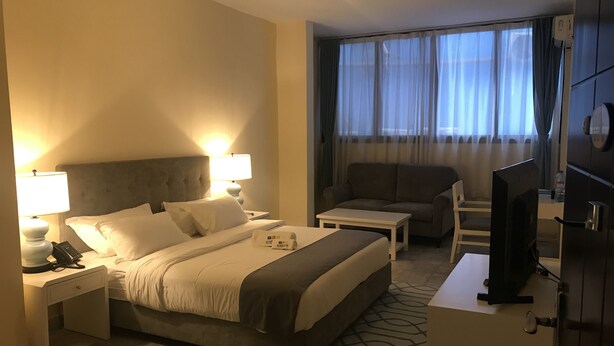 A hotel room with a bed, bedside lamps, a desk, a chair, a television, and a large window with curtains. - Sunset Inn Guest House (Monrovia)