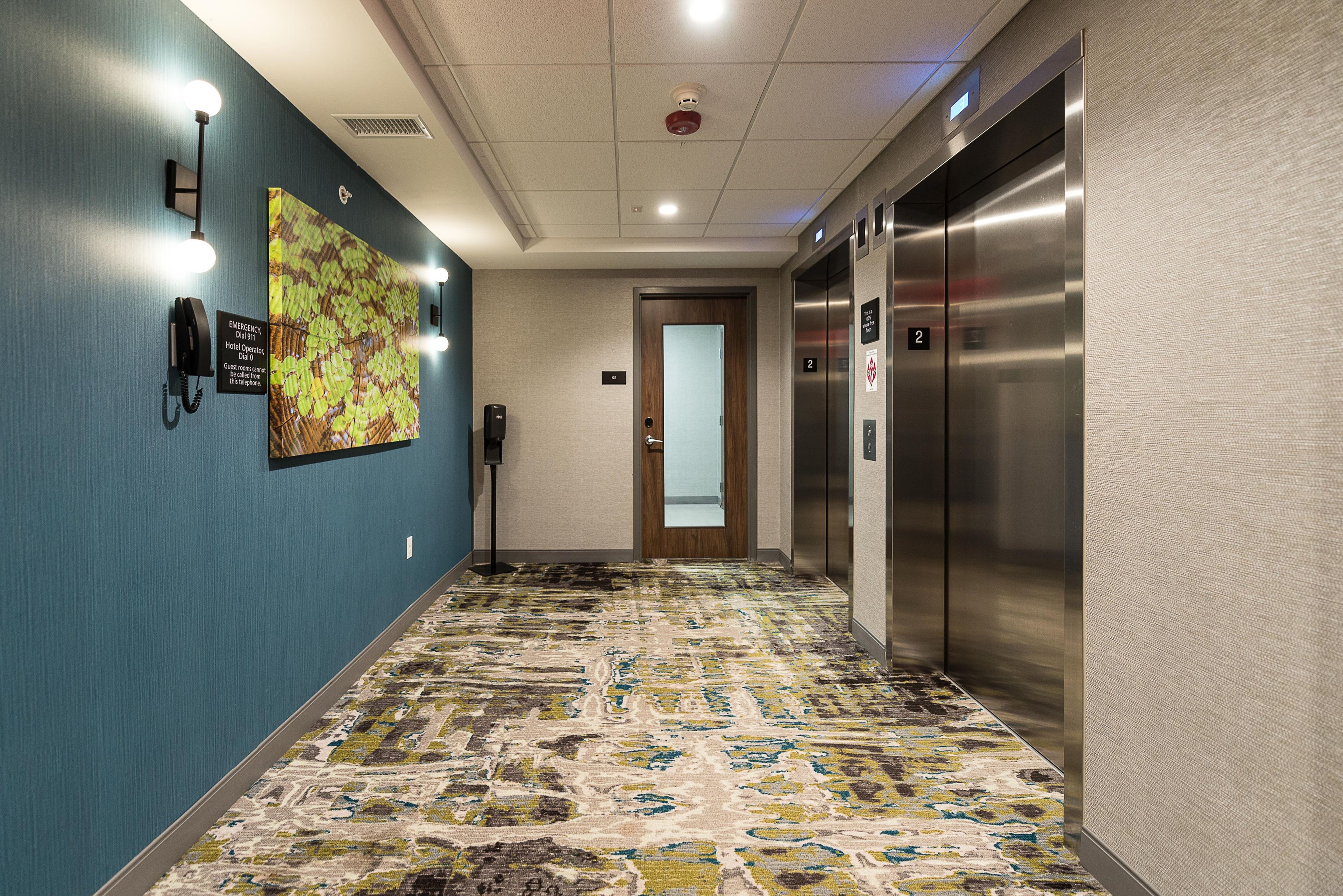 Een hotelgang met een patroon-tapijt, een noodtelefoon, een wandlamp en een lift.