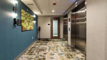 Een hotelgang met een patroon-tapijt, een noodtelefoon, een wandlamp en een lift.