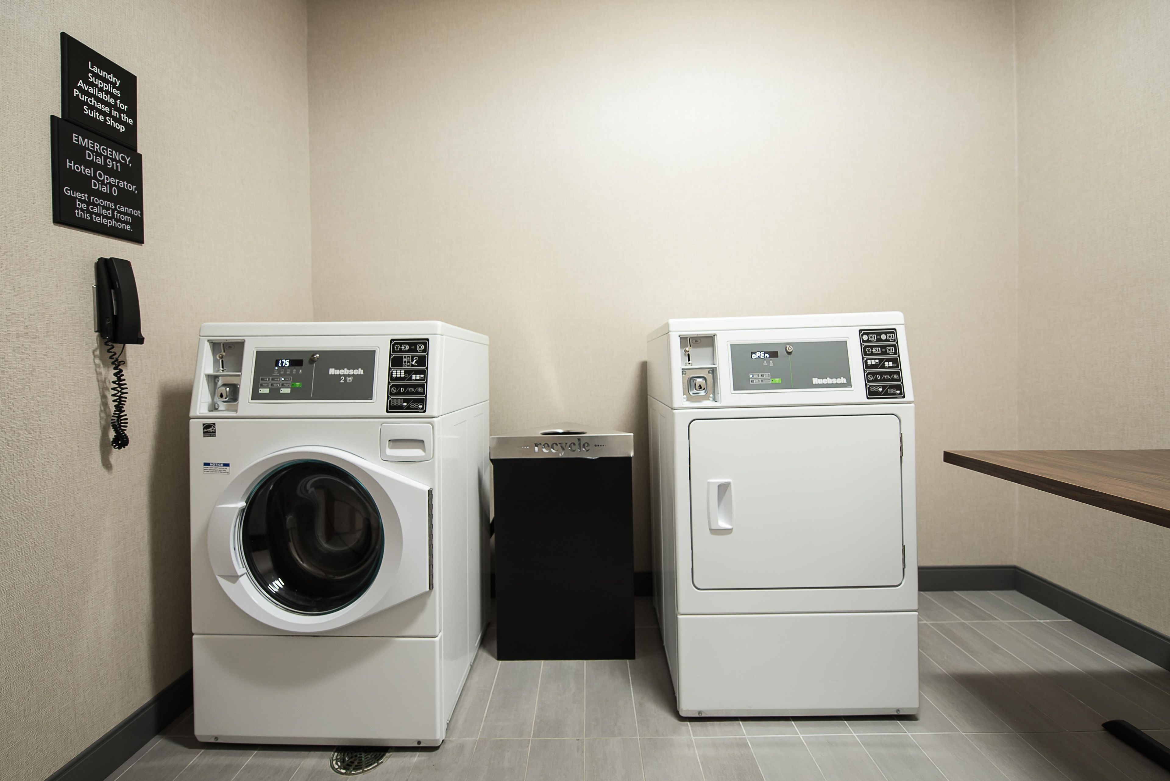 Zona de bugaderia amb dues rentadores i assecadores de càrrega frontal, un contenidor de reciclatge i un telèfon per assistència en cas d'emergència.
