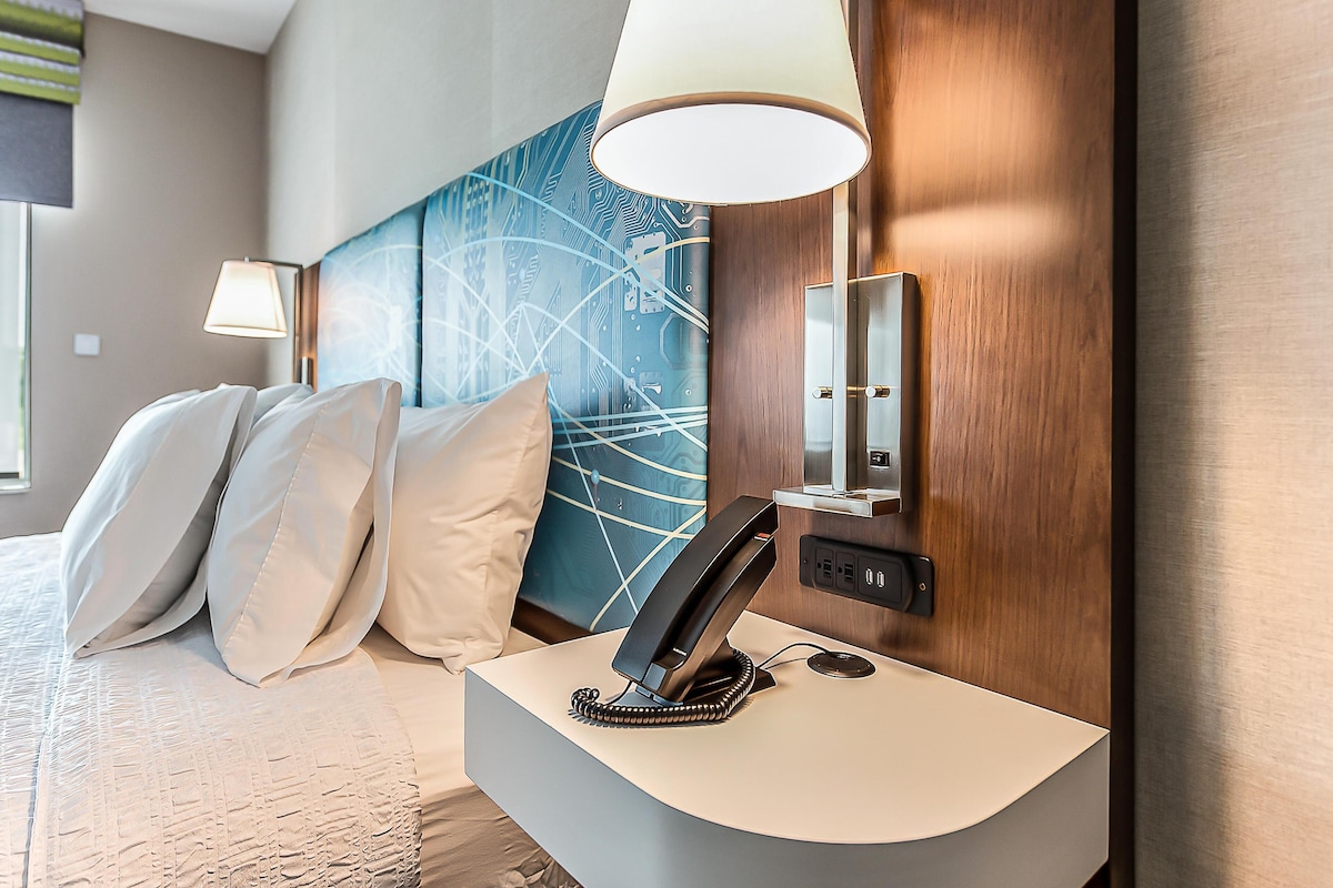 Una habitación de hotel moderna con una mesita de noche curvada, un teléfono, una lámpara fijada a la pared y un cuadro abstracto de gran tamaño.