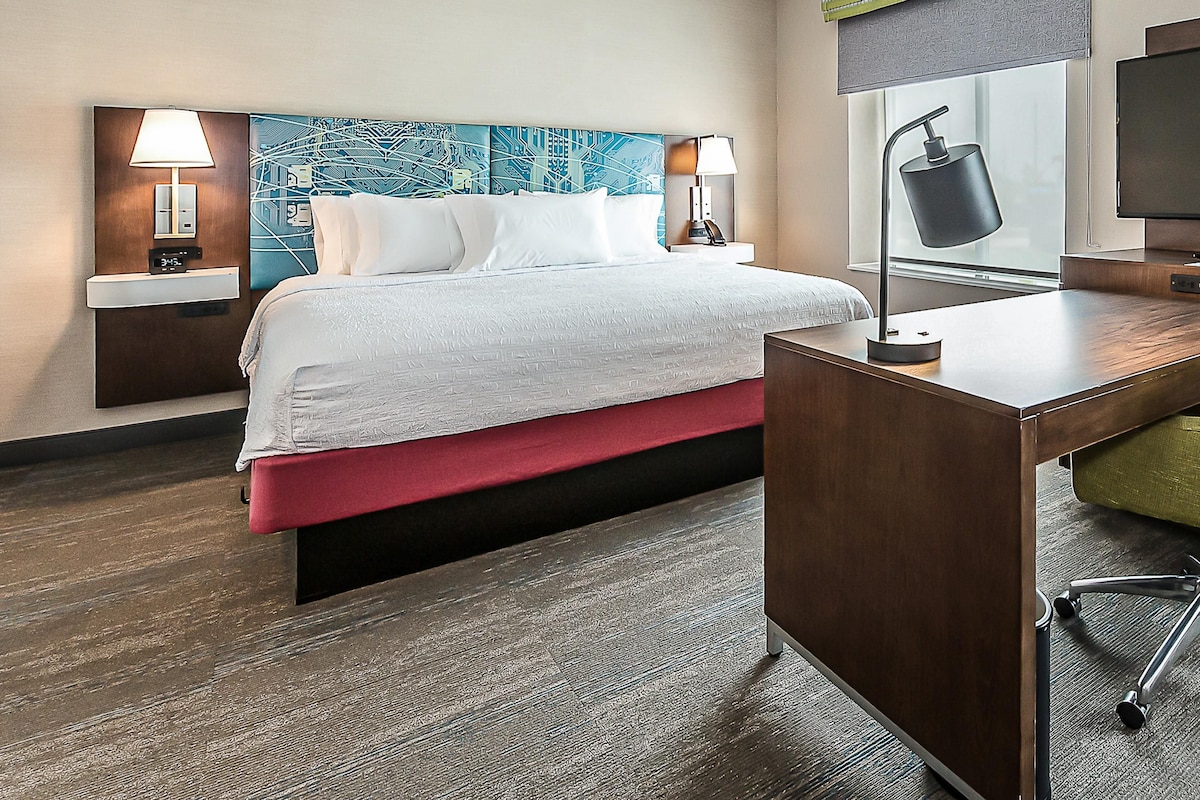 A hotel room with a bed, bedside tables, a desk, a chair, a TV, and a window with a view.
