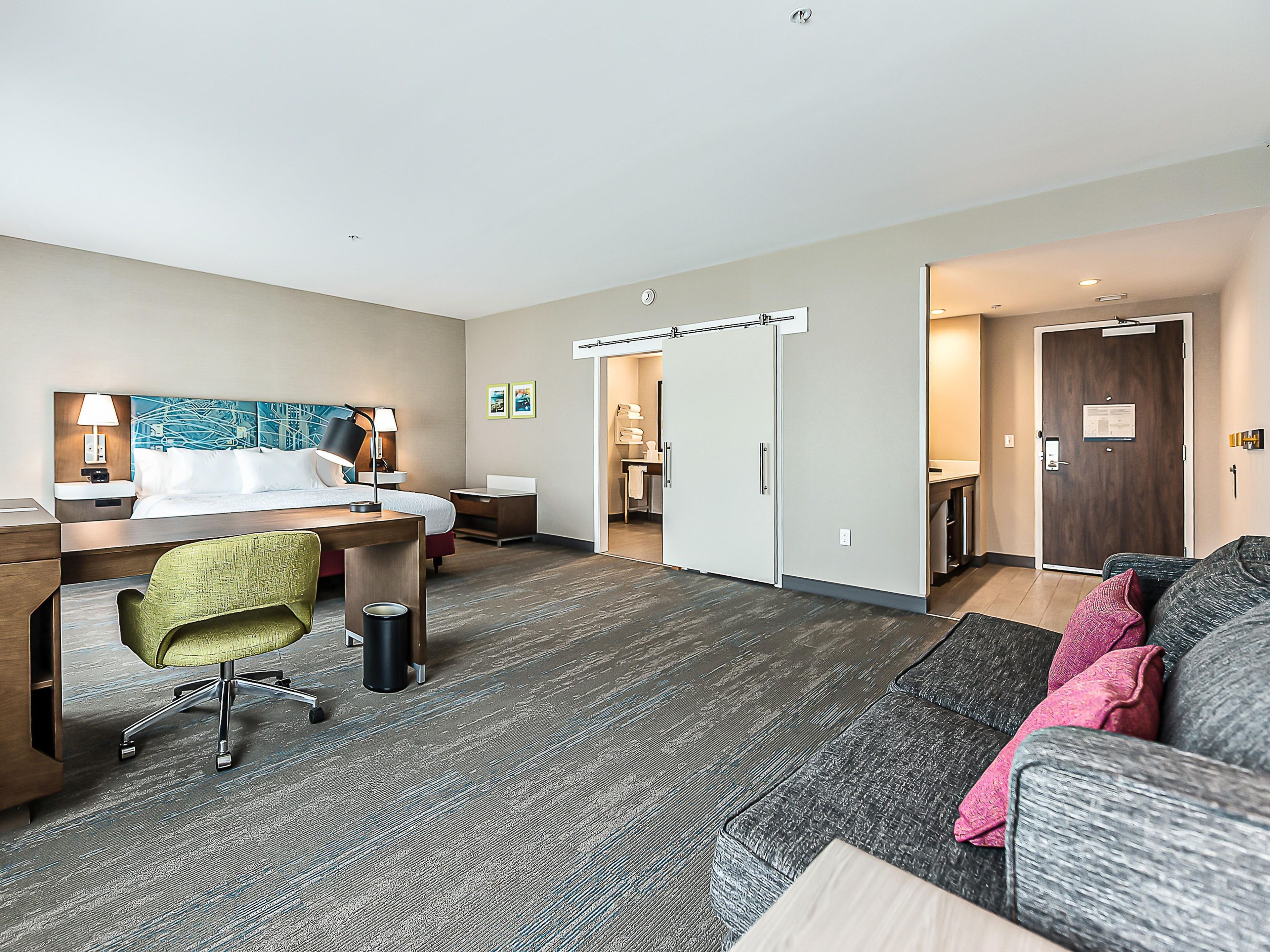 A hotel room with a bed, desk, chair, and a couch. There is a door leading to a bathroom and a hallway.