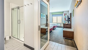 A modern hotel room with a glass shower enclosure, a bed with a headboard, a bedside table, and a desk with a chair.