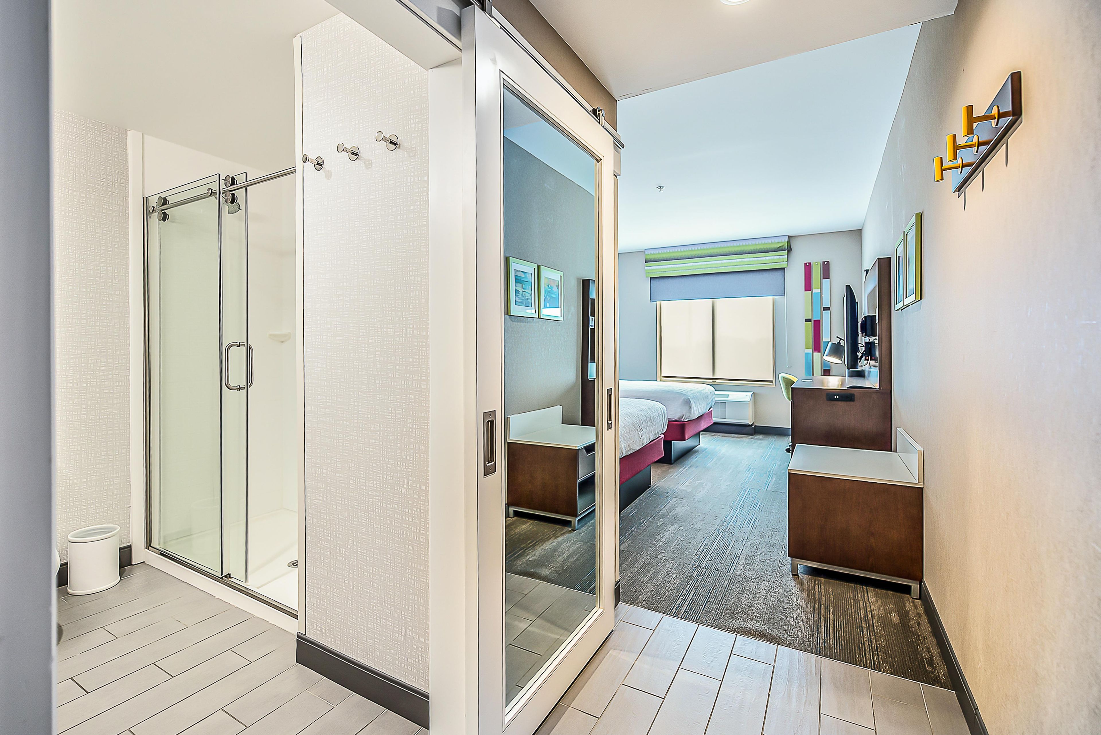 A modern hotel room with a glass shower enclosure, a bed with a headboard, a bedside table, and a desk with a chair.