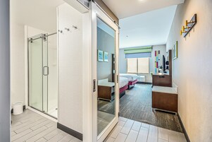 Habitación de hotel moderna con cabina de ducha de vidrio, cama con cabecero, mesita de noche y escritorio con silla.