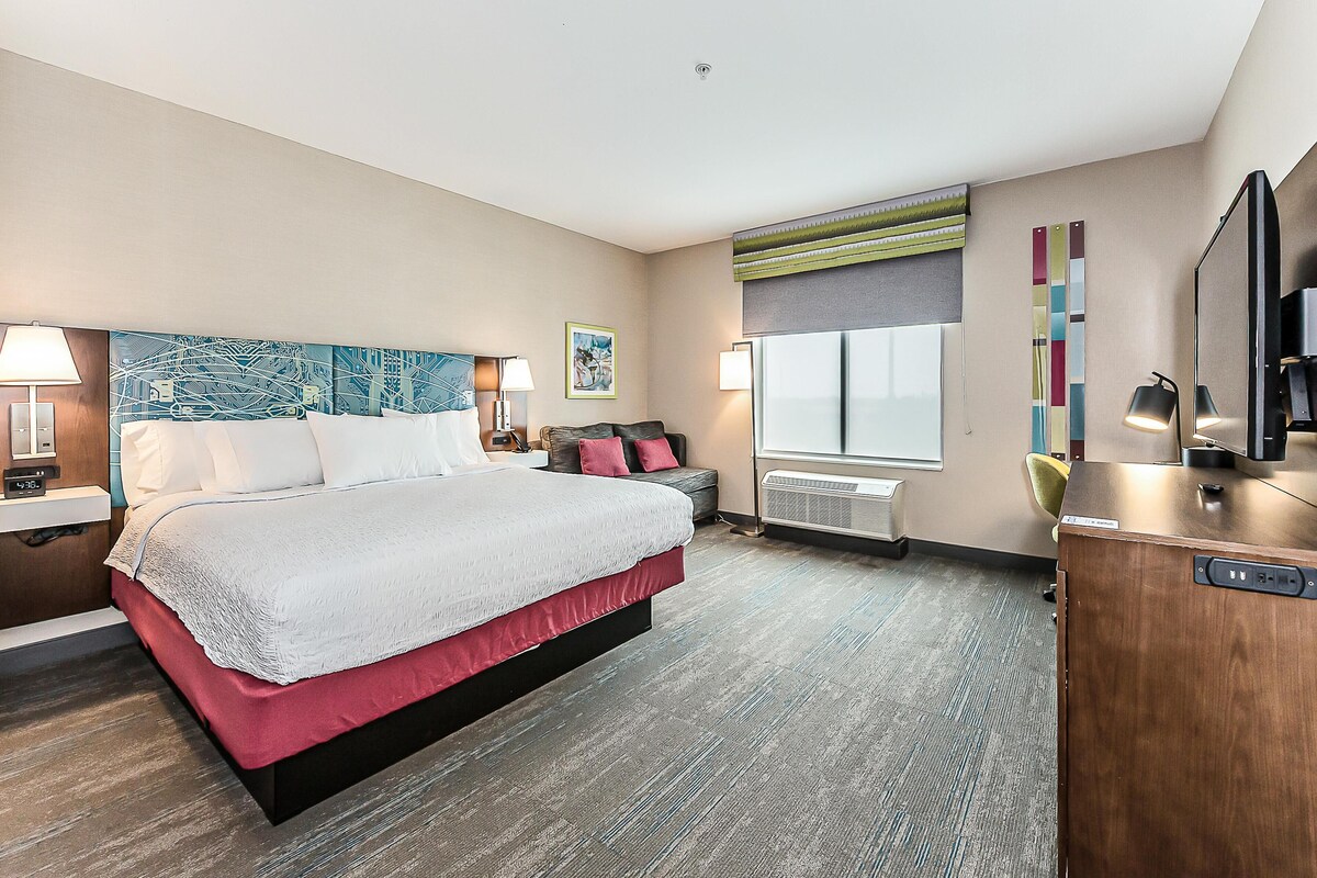 Habitación de hotel con una cama grande, un escritorio, una silla, un televisor y una ventana con persianas.