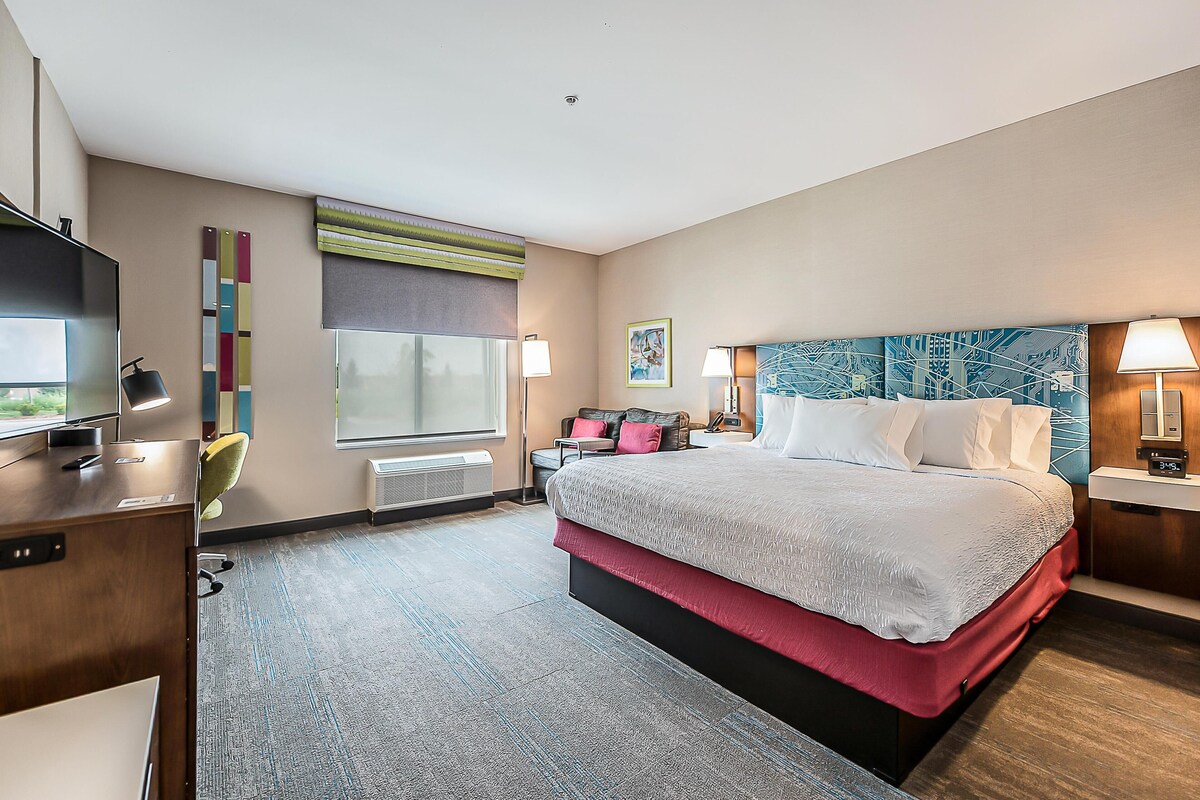 Habitación de hotel con una cama grande, un escritorio con televisor, una ventana con persianas y un mural de un paisaje urbano en la pared.