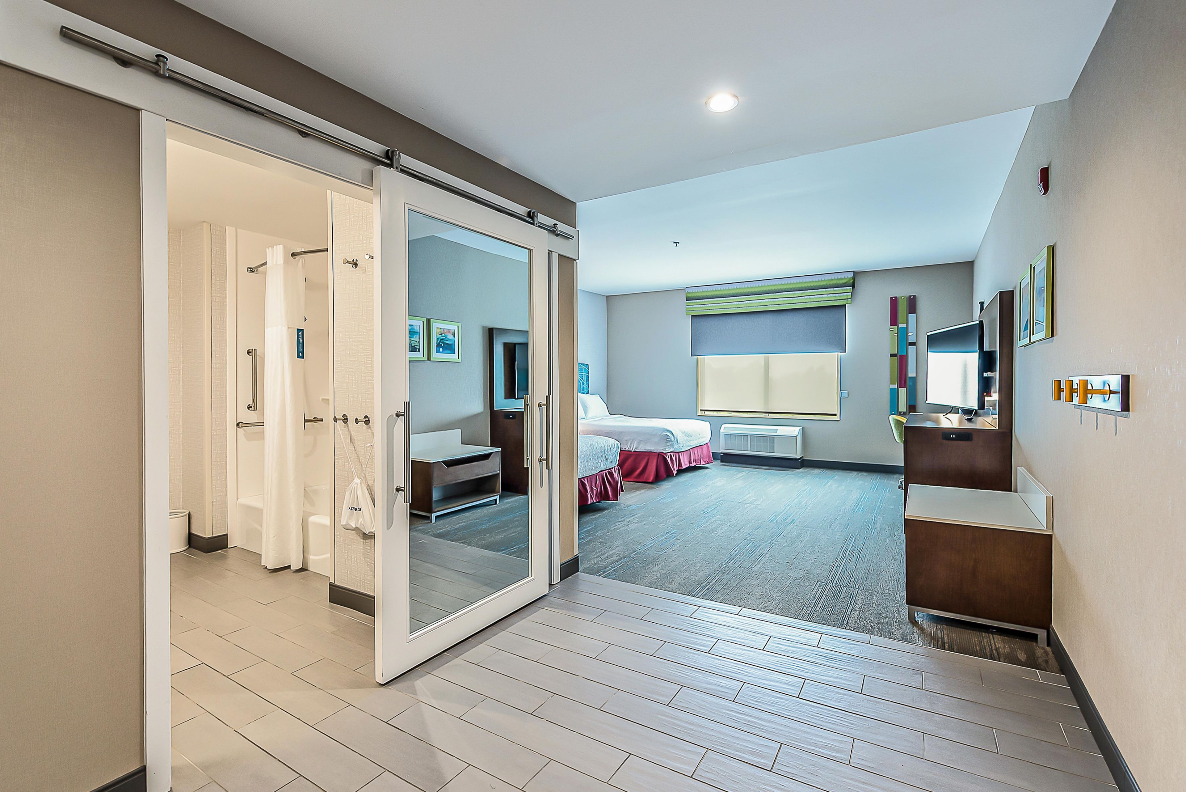 Una habitación de hotel moderna con una puerta corredera que da a un baño, una cama con falda roja, un escritorio y un ventanal grande.