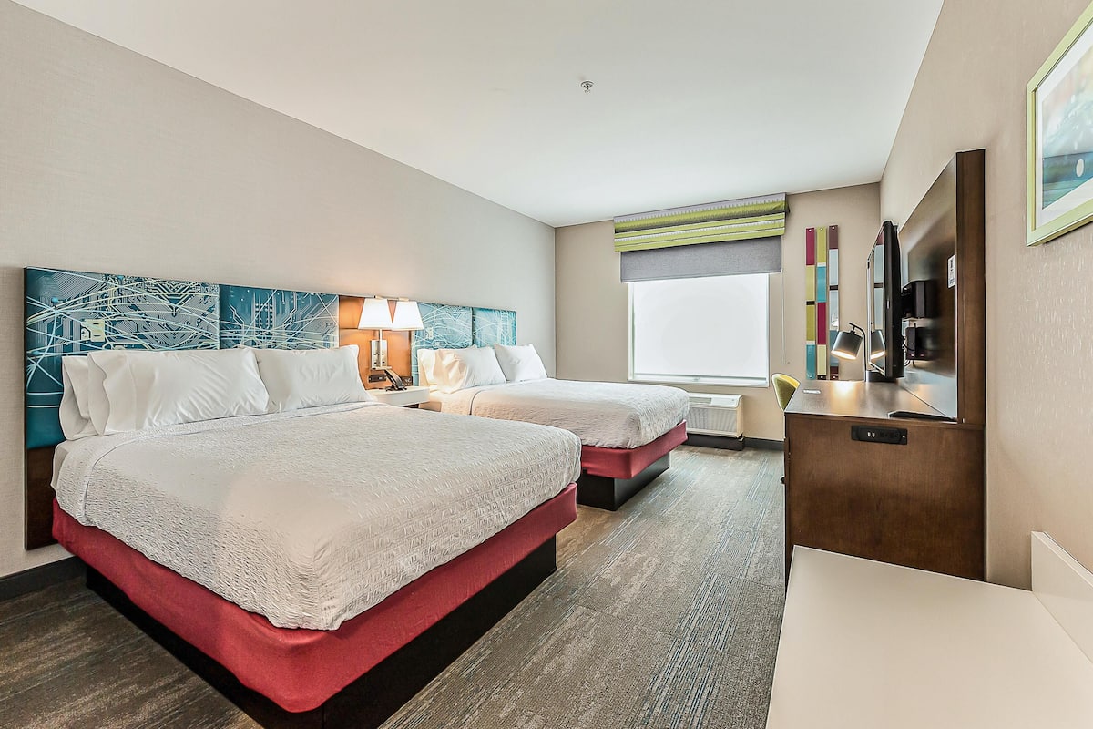 Quarto de hotel com duas camas, uma escrivaninha, uma cadeira, uma televisão e uma janela grande.