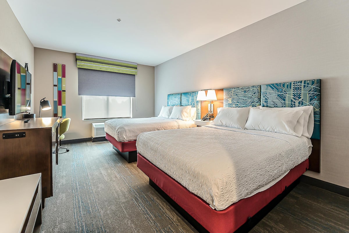 Quarto de hotel com duas camas, uma escrivaninha, uma cadeira e uma janela grande com persianas.