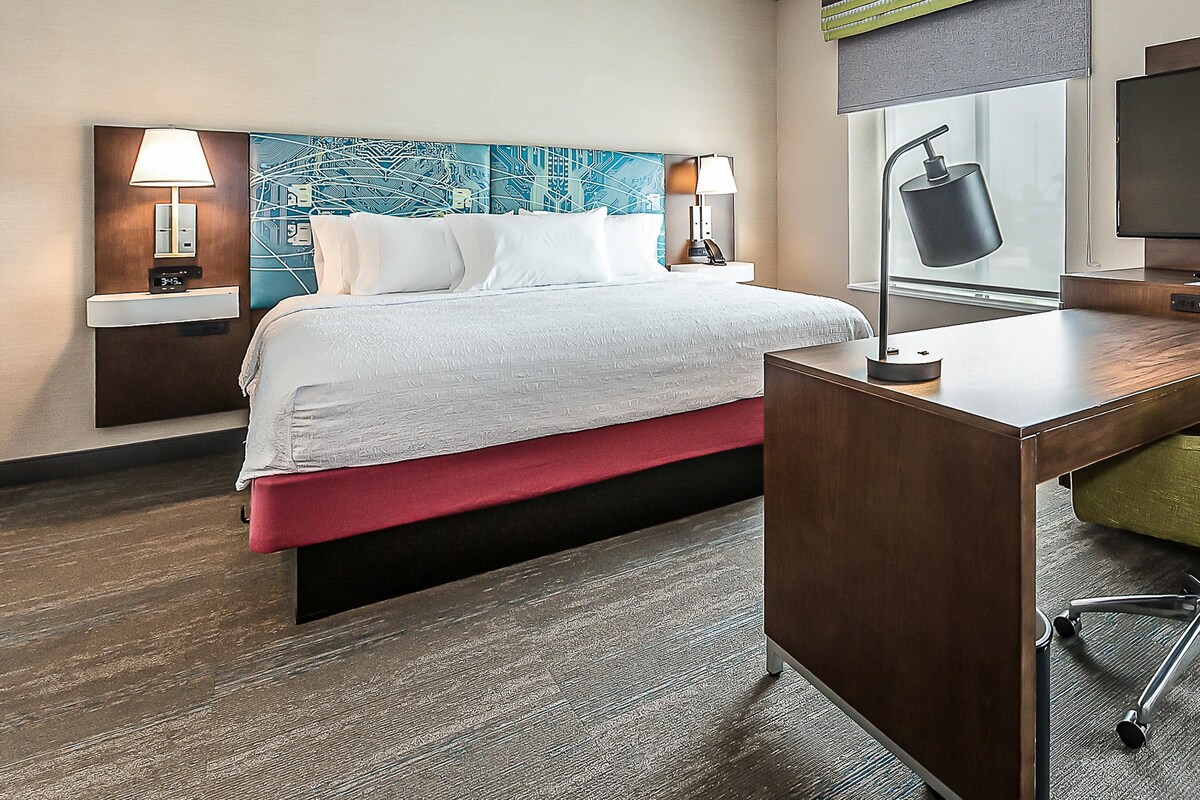 Una habitación de hotel con cama, mesitas de noche, escritorio, silla, televisor y ventana con vistas.
