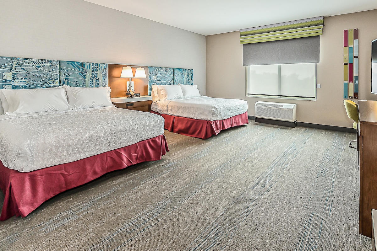 A hotel room with two beds, a nightstand with a lamp, a window with blinds, and a wall-mounted air conditioner.