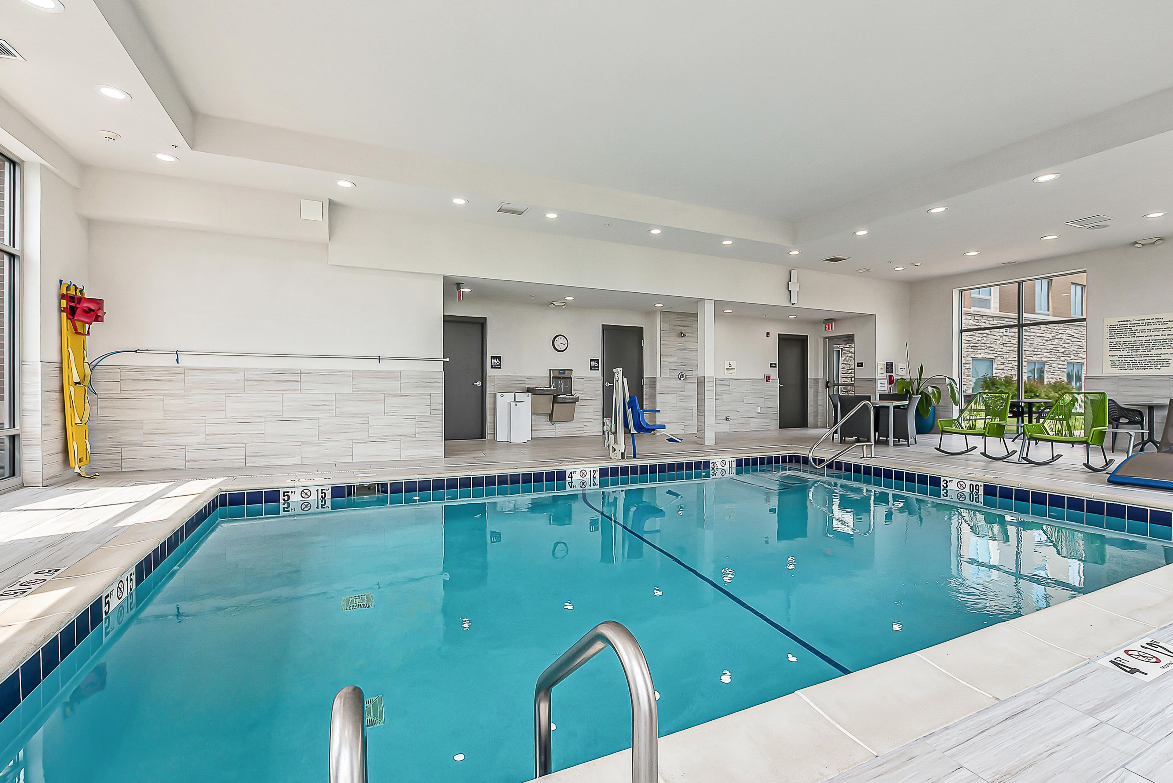 A modern indoor swimming pool with clear lane markings, a lifeguard chair, and a reception area with a clock and informational posters.