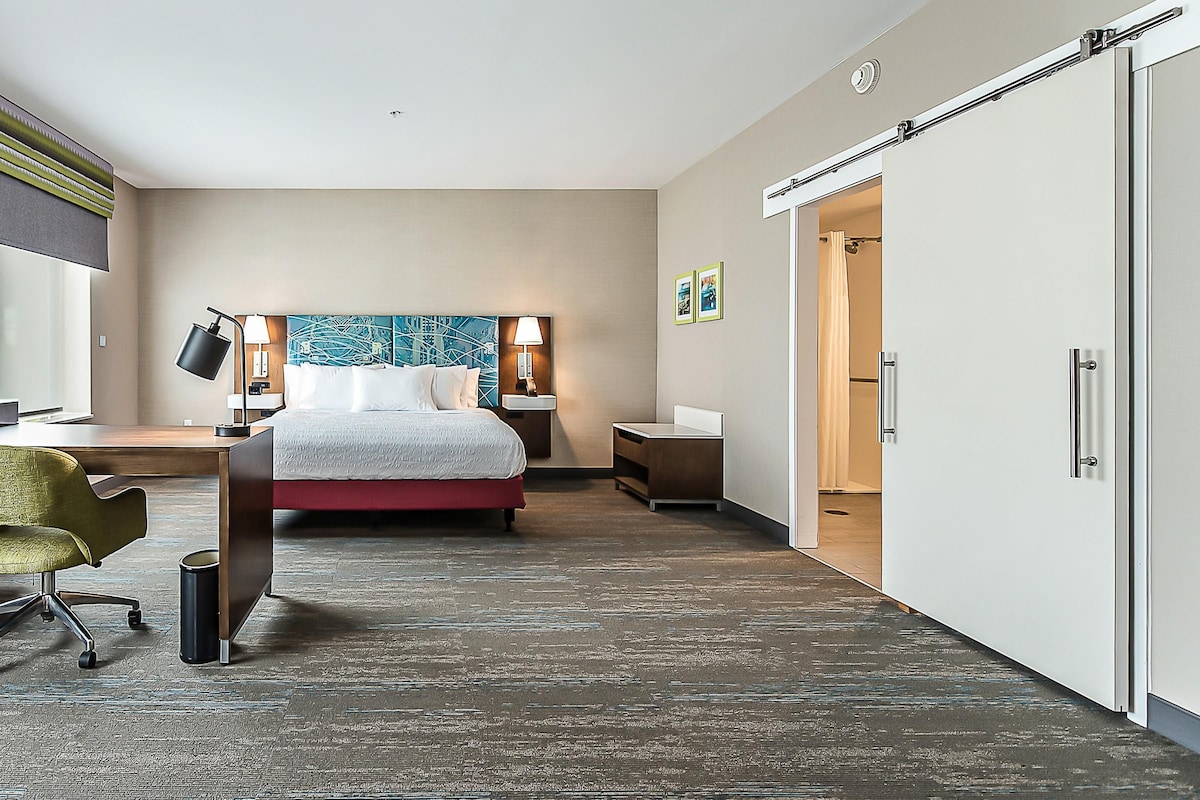 A modern hotel room with a large bed, a desk with a chair, a wardrobe, and a bathroom visible through an open door.