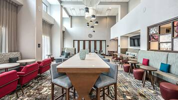 A hotel lobby with a long communal table, red and grey seating, and a patterned carpet.