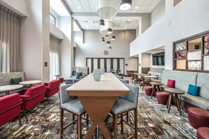 A hotel lobby with a long communal table, red and grey seating, and a patterned carpet. - Hampton Inn & Suites Howell (Howell)