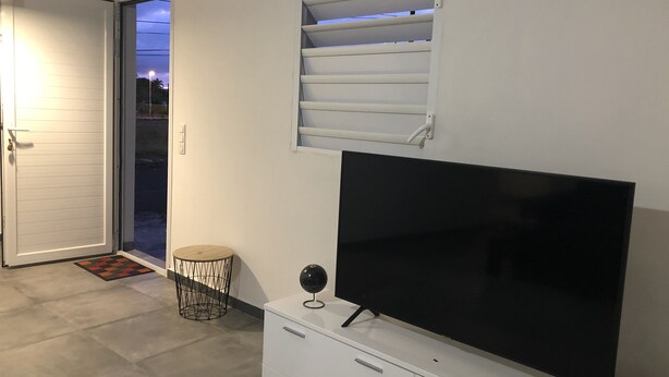 Ein modernes Wohnzimmer mit einem Flachbildfernseher auf einer weißen Kommode, einem kleinen runden Hocker und einem Fenster mit Jalousien.