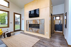 A modern living room with a large TV, a rustic fireplace, and a view of the outside. - Lake + Ledge Retreat l Modern Luxury Home on Pike Lake (Hatley)