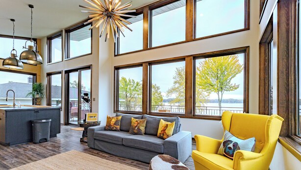 A spacious living area with a gray sofa, a yellow armchair, and a large window overlooking a tree. - Lake + Ledge Retreat l Modern Luxury Home on Pike Lake (Hatley)