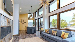 A modern living room with a grey sofa, a large window, and a kitchen area. - Lake + Ledge Retreat l Modern Luxury Home on Pike Lake (Hatley)