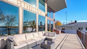 A modern outdoor deck with a wicker sofa and a coffee table, overlooking a body of water and a distant building. - Lake + Ledge Retreat l Modern Luxury Home on Pike Lake (Hatley)