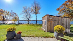 A wooden shed with a metal door, surrounded by grass and trees, near a body of water. - Lake + Ledge Retreat l Modern Luxury Home on Pike Lake (Hatley)