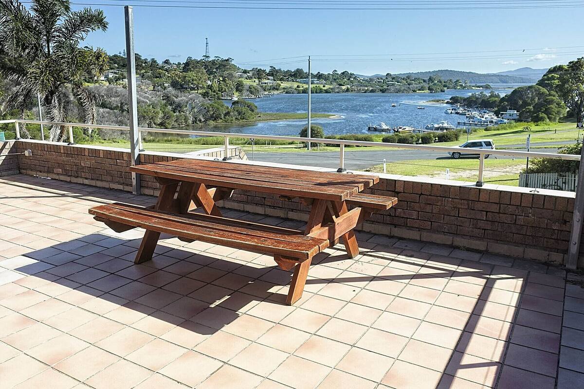 A wooden picnic table on a tiled patio with a view of a lake and boats.