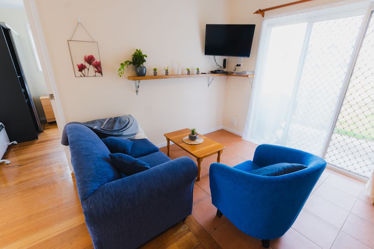 A living room with a blue sofa, a blue armchair, a small wooden table, a TV mounted on the wall, and a plant on a shelf.