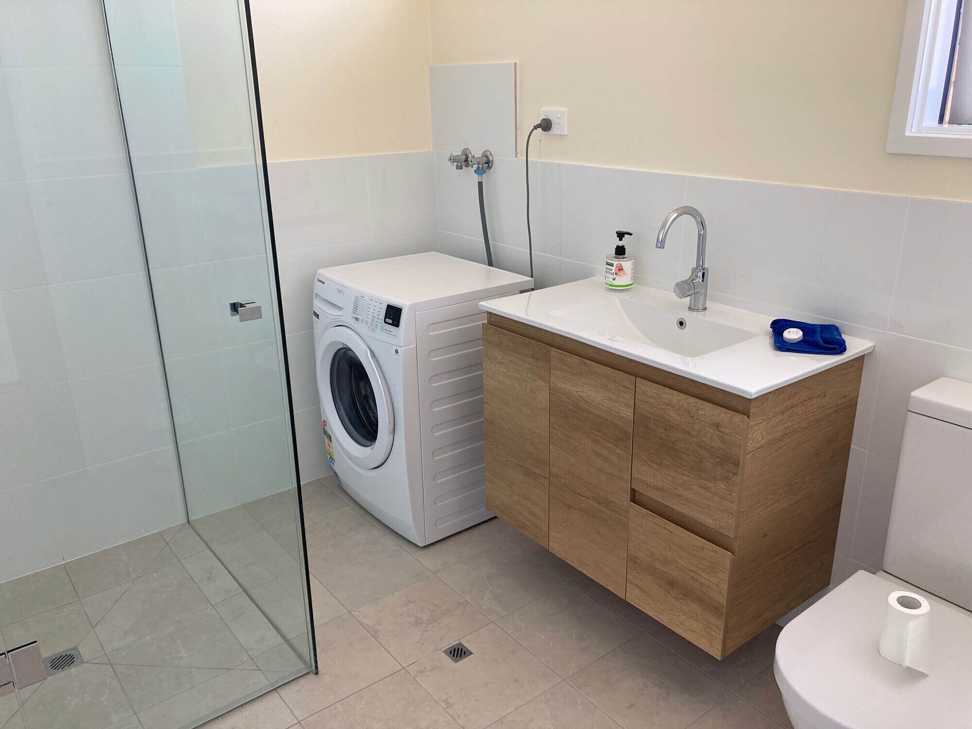 A modern bathroom with a glass shower enclosure, a built-in washing machine, a wooden vanity with a sink, and a toilet.