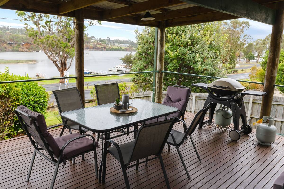 A covered outdoor dining area with a table and chairs, overlooking a lake with boats.
