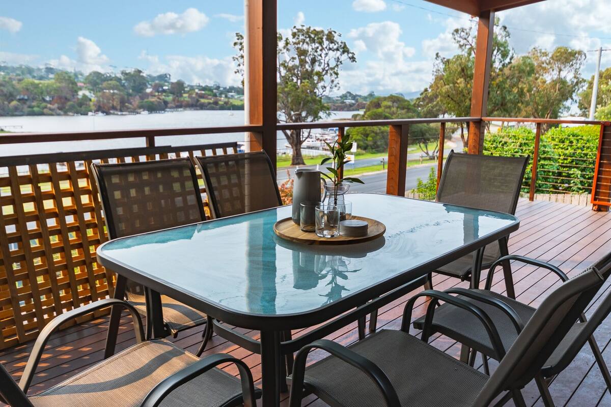 A glass-top table with four chairs, a pitcher, and a small plant on a wooden deck overlooking a body of water.