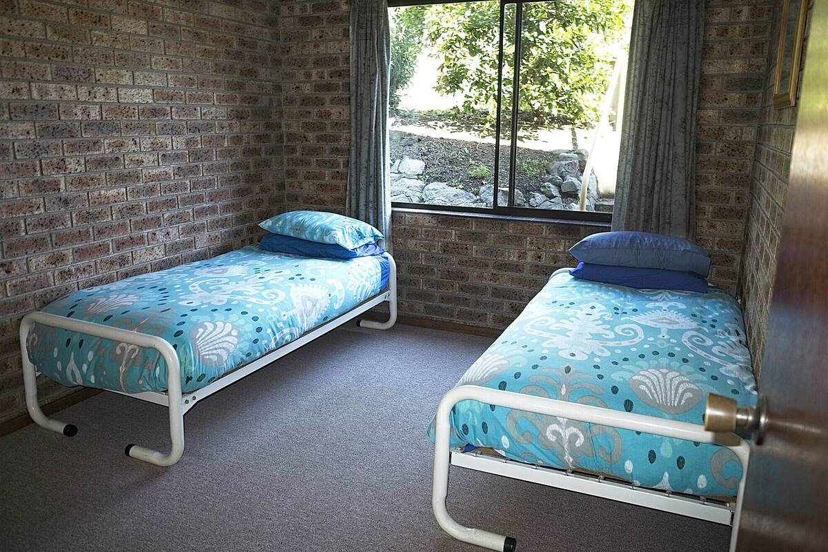 Two single beds with patterned blue bedding in a room with brick walls and a window with a view of trees and rocks.