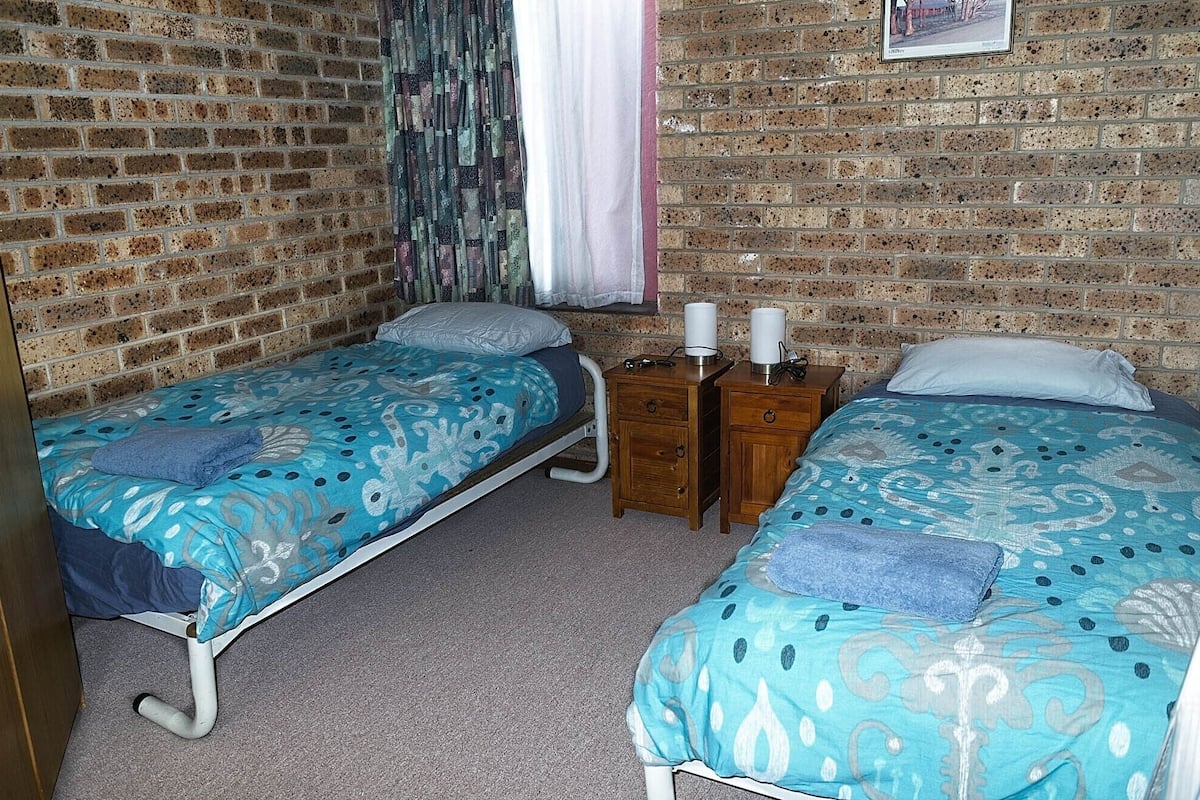 A room with two single beds, each with a patterned blue bedspread and pillows, wooden nightstands with lamps, and a brick wall.