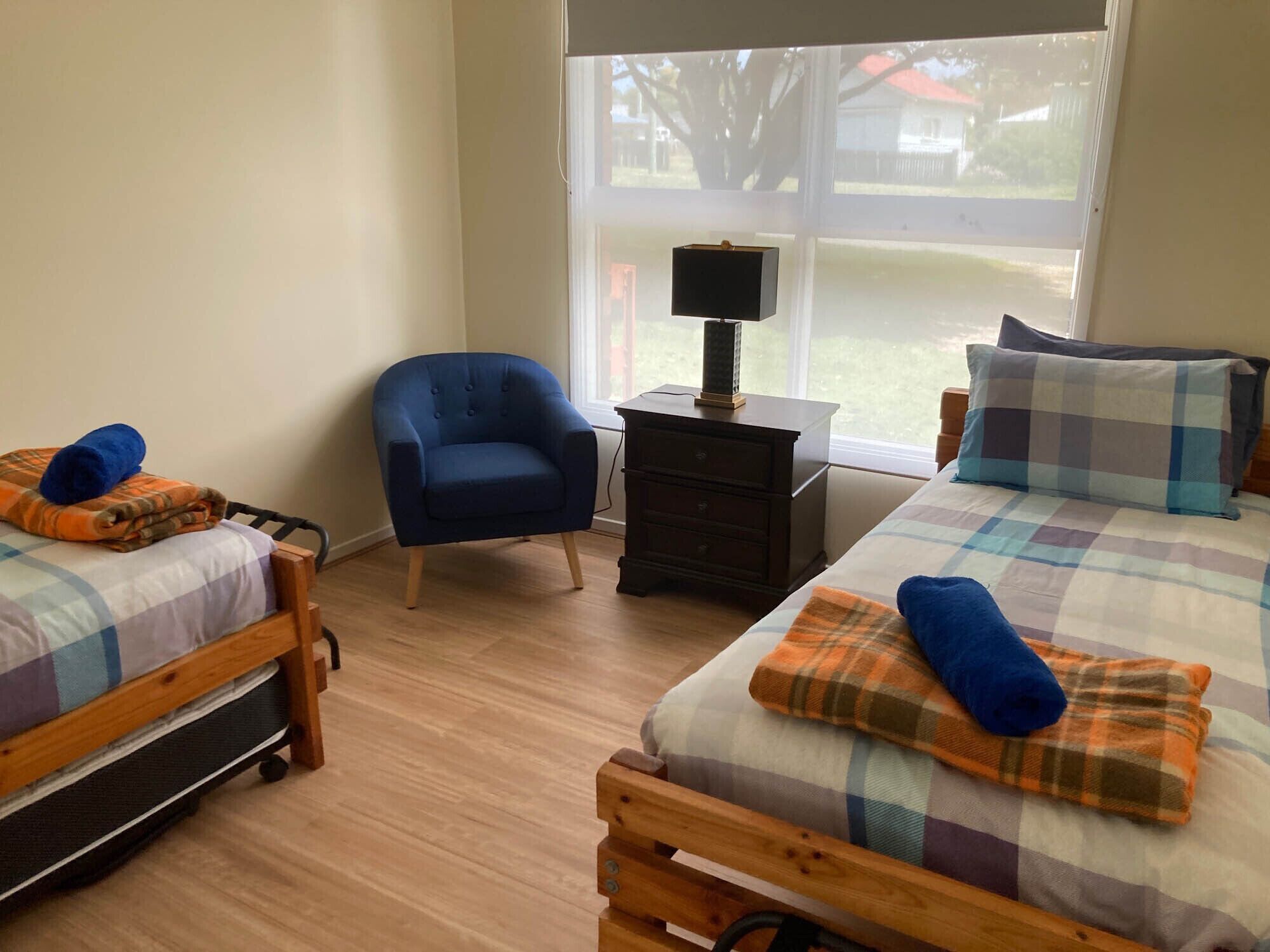 A bedroom with two wooden beds, a blue armchair, a nightstand with a lamp, and a window with a view of the outdoors.