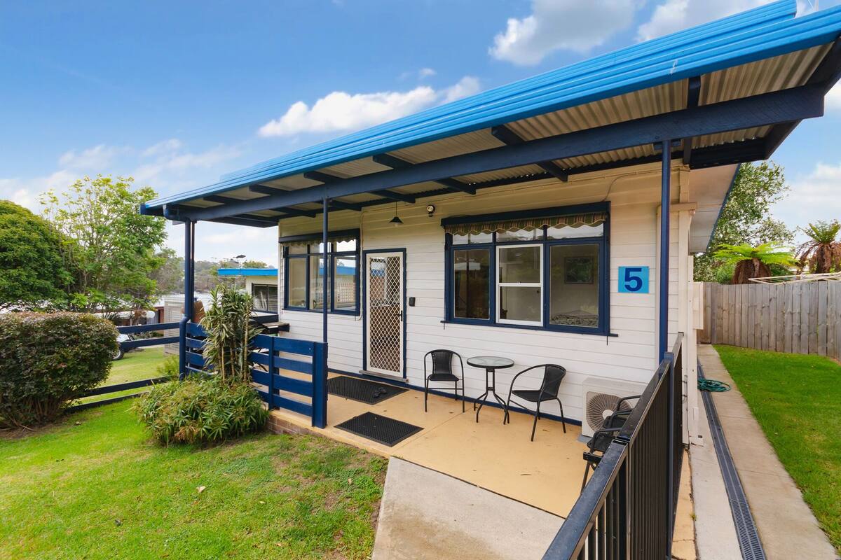 A single-story building with a blue roof and white walls, number 5, featuring a covered patio with two chairs and a table, surrounded by a small lawn and a wooden fence.
