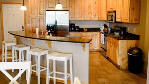 Una cocina con armarios de madera, un refrigerador de acero inoxidable, un microondas y un lavamanos. Hay tres taburetes blancos y una encimera con una cafetera y otros electrodomésticos.