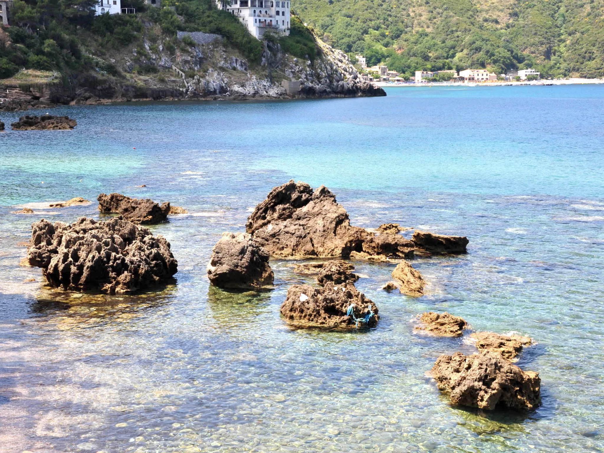 A coastal area with clear water, rocky formations, and buildings on a hillside.