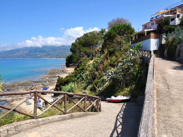 A coastal road with a wooden fence, a boat, and a beach with umbrellas.